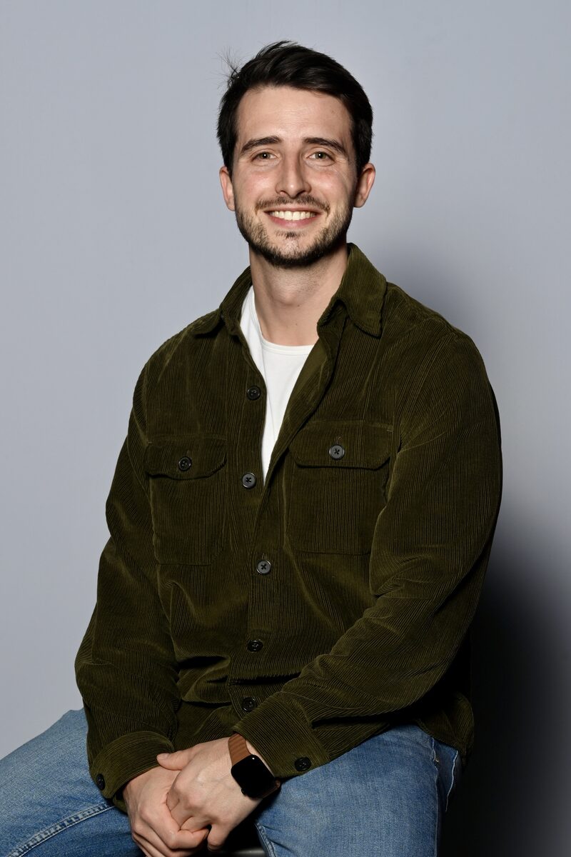 Retrato de estudio de Joan Boluda Chova, sentado y sonriendo, con sobrecamisa verde de pana sobre fondo gris.